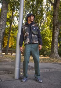 Fleece vest with camouflage pattern, black hoodie, green pants, grey sneakers. Standing by a lamp post in a forested area.