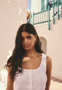 White textured tank top with a scoop neckline and three buttons. Model has long dark hair and wears gold hoop earrings. Bright backdrop.
