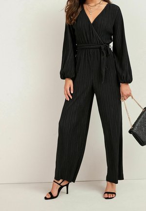 Black striped jumpsuit with long, puffed sleeves, a wrap design, and a tied waist. Worn with black strappy heels and a quilted handbag.