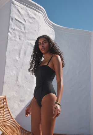 Frau mit lockigem Haar, die einen schwarzen Einteiler trägt, steht neben einer gebogenen weißen strukturierten Wand unter einem klaren blauen Himmel.