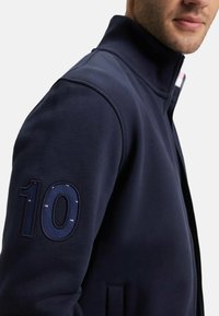 Sweatshirt zippé marine avec col haut, texture douce et un grand "10" brodé sur la manche gauche. Inclut une fermeture éclair cachée.
