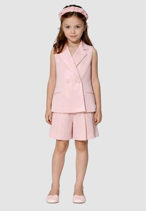 Ragazza giovane in piedi, indossa un gilet doppiopetto rosa senza maniche, pantaloncini plissettati abbinati, una fascia rosa per capelli e scarpe rosa con fiocchi.