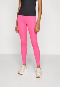 Sitter, høy midje leggings i lys rosa, laget av stretchy materiale, kombinert med lette joggesko. Har en subtil logo nær ankelen.