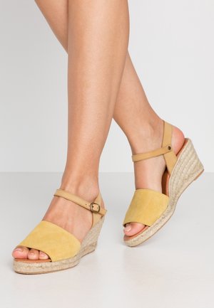Jambes portant des sandales compensées en daim jaune avec des brides à la cheville et des talons plateforme en jute tressé sur un fond clair uni.