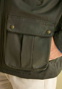 Ciemnozielona kurtka z zasuwaną kieszenią z klapką, gładką fakturą i detalami szwów. Logo widoczne na kieszeni.