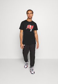 T-shirt noir arborant un logo des Tampa Bay Buccaneers en rouge et blanc. Le modèle porte un jogging noir et des baskets blanches avec des accents rouges.