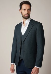 Mężczyzna w ciemno teksturowanej marynarce, pasującym kamizelce, białej koszuli i ciemnych spodniach, stojący na tle jasnym, patrzący w bok.