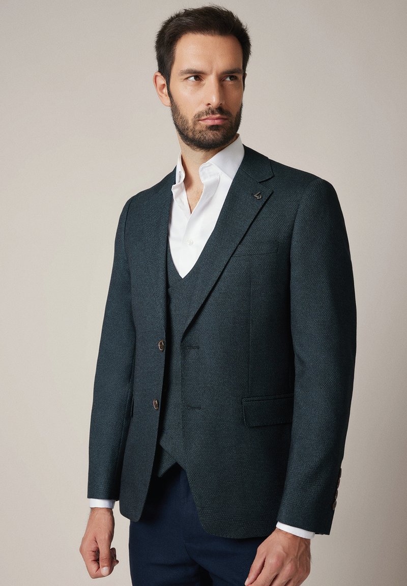 Mężczyzna w ciemno teksturowanej marynarce, pasującym kamizelce, białej koszuli i ciemnych spodniach, stojący na tle jasnym, patrzący w bok.