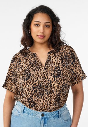 Mladá žena s vlnitými tmavými vlasmi, ktorá má na sebe blúzku s krátkym rukávom s leopardím vzorom a svetlomodré džínsy s vysokým pásom.