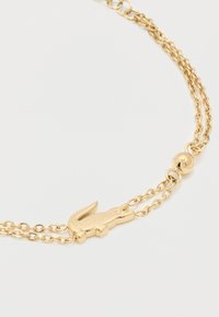 Χρυσό βραχιόλι με διπλή αλυσίδα και κεντρικό φυλαχτό σε σχήμα κροκόδειλου σε απλό λευκό φόντο.