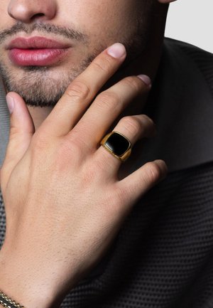Goldring mit einem quadratischen schwarzen Stein, getragen an einer Hand mit heller Haut, die eine glatte Oberfläche und einen polierten Glanz zeigt.