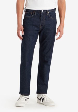 Hombre vistiendo jeans de pierna recta de color azul oscuro y zapatillas blancas frente a un fondo blanco liso.