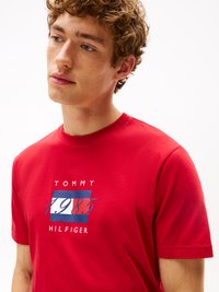Tricou din bumbac roșu cu un design grafic pe față, având inscripțiile "TOMMY" și "1985" în albastru și alb. Mâneci scurte și guler rotund.