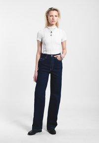 Bílý přiléhavý tričko s krátkými rukávy, tmavě modré vysoké široké džíny, černé boty a vrstvené stříbrné náhrdelníky.