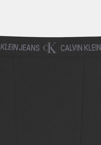 Calças pretas Calvin Klein Jeans com uma cintura elástica larga, apresentando o texto "CALVIN KLEIN JEANS" e o logo "CK" em texto cinza. Textura suave, design ajustado.