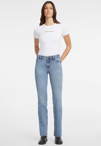 Bela prilegajoča majica s kratkimi rokavi s logotipom "GUESS JEANS", v kombinaciji s svetlo modrimi ravnimi hlačami, minimalistična zasnova, gladka tekstura in črne škornje.