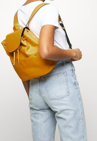 Persona con camiseta blanca y jeans azul claro lleva una mochila de cuero amarillo mostaza sobre un hombro.