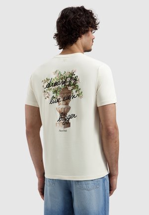 Man met krullend haar die een wit T-shirt draagt met een ontwerp van een bloemenvaas en de tekst "droom groot, leef nog groter" op de achterkant.