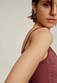 Camiseta de tirantes finos en color burdeos, con un diseño ajustado y costuras visibles. Textura de tela sencilla y suave. Pendientes geométricos.