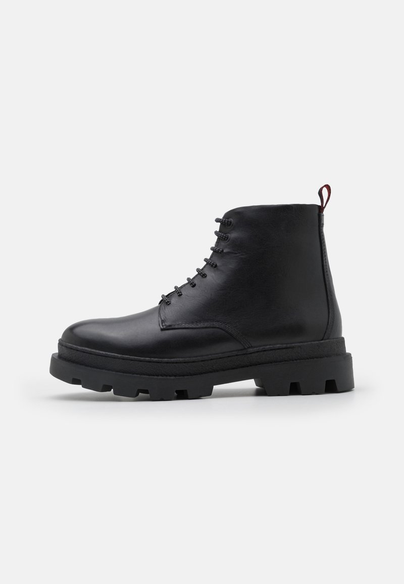 Bottine noire en cuir à lacets avec semelle crantée épaisse et languette de tirage rouge sur fond blanc.