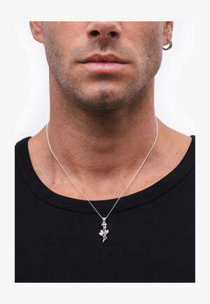 Ezüst medál nyaklánc virágmintával, vékony láncon lógva, fekete bordás póló felett viselve.