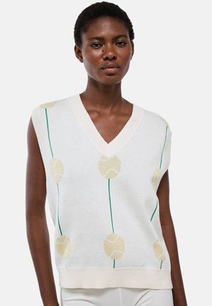 Femme portant un gilet en maille blanc sans manches avec des motifs beige de balles de tennis et des tiges vertes, associé à un pantalon blanc, sur un fond uni.
