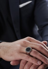 Sølvring med en glatt svart oval stein, satt i et polert metallbånd, hviler på en hånd mot en bakgrunn av mørk dressjakke.