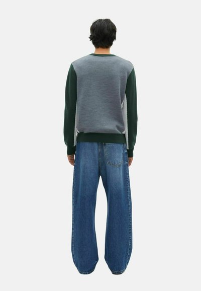 Persona in piedi di spalle che indossa un maglione grigio e verde con jeans blu larghi su uno sfondo semplice.