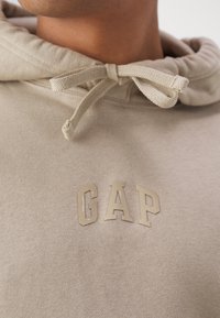 Krémová mikina s kapucí a šňůrkou, na které je texturované logo "GAP" v světlejším odstínu. Měkký bavlněný materiál a uvolněný střih.