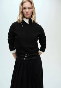 Maglione nero a maglia con colletto alto, abbinato a una gonna plissè nera con due cinture con fibbie. I polsini bianchi della camicia sono visibili.