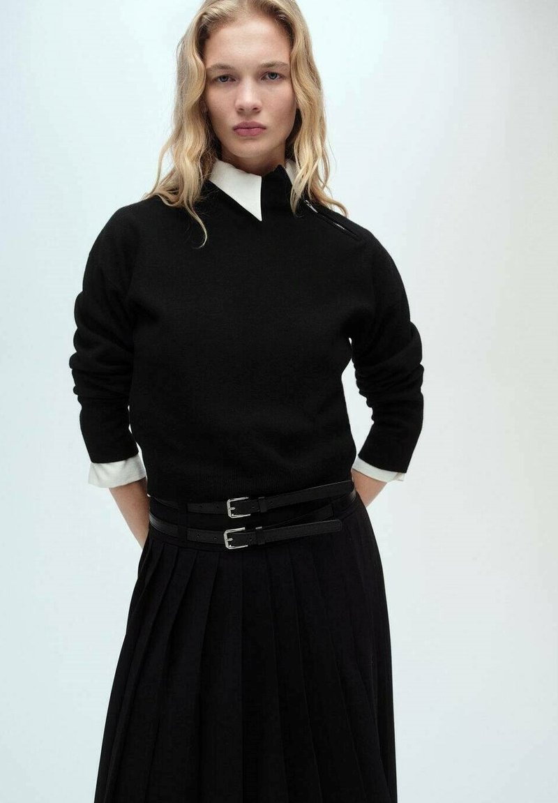 Maglione nero a maglia con colletto alto, abbinato a una gonna plissè nera con due cinture con fibbie. I polsini bianchi della camicia sono visibili.