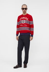 Homme portant des lunettes de soleil noires, un pull rayé rouge et marine avec des inscriptions blanches, un pantalon noir et des chaussures marron foncé, debout sur un fond blanc.
