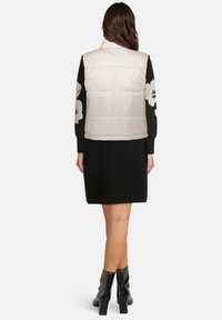 Jasnobeżowa kamizelka puchowa z wysokim kołnierzem, o gładkiej fakturze i pikowanym wzorze, noszona na czarnej sukience z kwiatowymi akcentami.