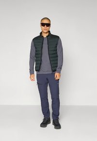 Grønn polstret vest over en grå langermet skjorte, kombinert med mørkeblå cargobukser og svarte tursko. Tilbehør inkluderer solbriller.