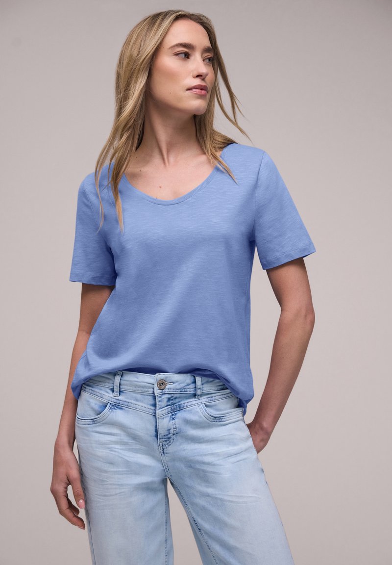 Mujer con cabello largo y rubio que lleva una camisa de manga corta azul claro y jeans de lavado claro, de pie con una mano en el bolsillo, mirando hacia la izquierda.