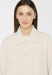 Helle beige übergroße Jacke mit Kragen, zwei Brusttaschen und vier Knöpfen. Hergestellt aus einem strukturierten Stoff für zusätzliche Details.