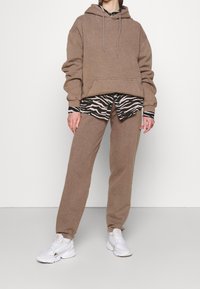 Brązowa, oversize bluza z kapturem ze ściągaczami, zestawiona z koszulą w zebra wzór oraz dopasowanymi brązowymi dresami. Uzupełnione białymi sneakersami.