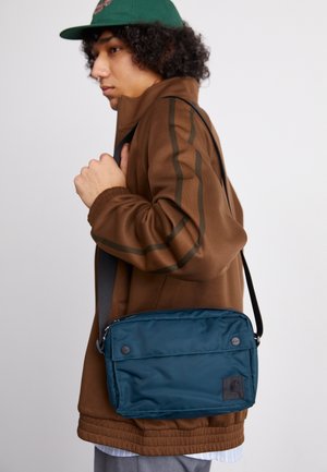 Personne portant une veste marron et une casquette verte porte un sac bandoulière bleu avec une bandoulière noire sur l'épaule.