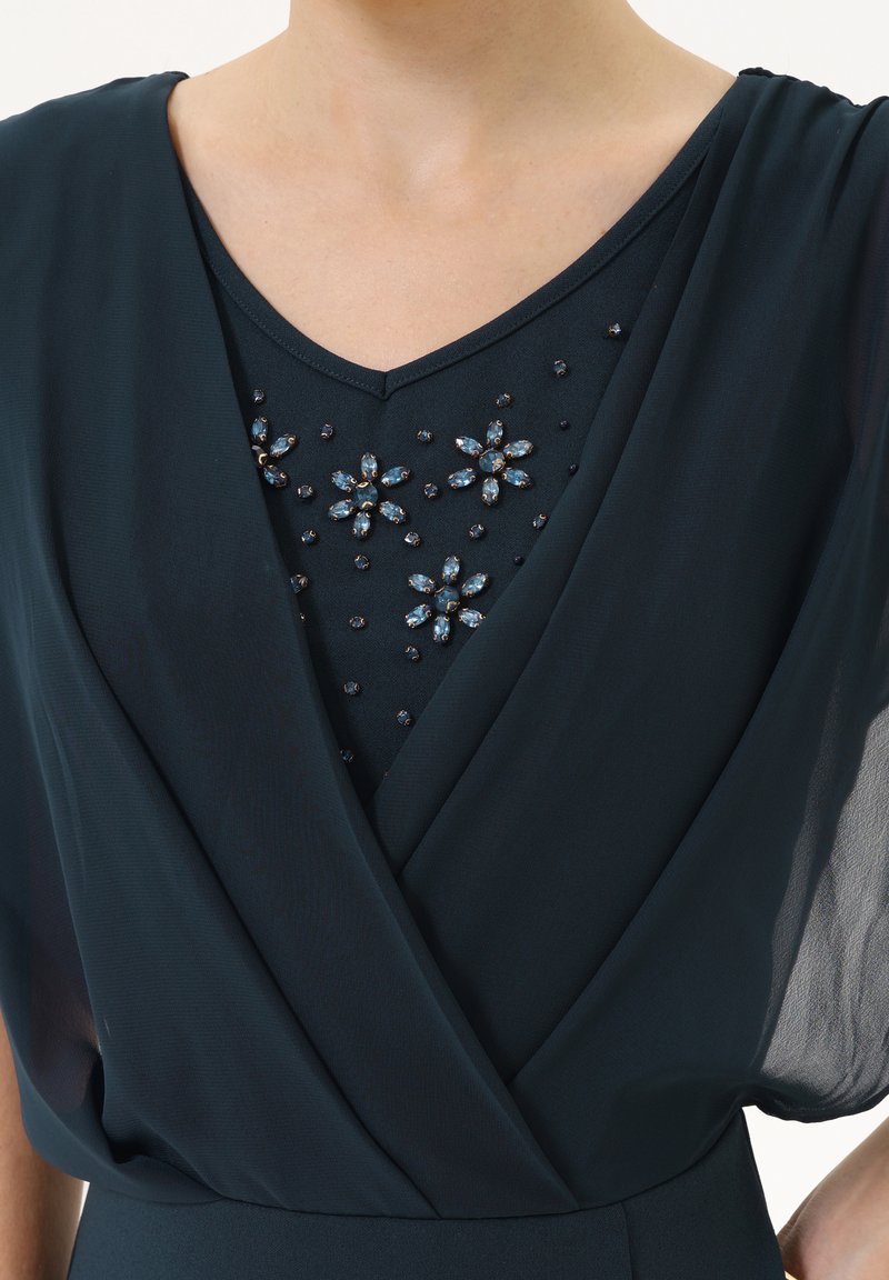 Jurk van donkerteal stof heeft een v-halslijn met bloemige versieringen van blauwe edelstenen en zwarte kralen, en sheer gedrapeerde mouwen.