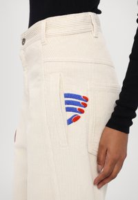 Bílé manšestrové kalhoty se boční kapsou, zdobené vyšitými modrými a červenými akcenty. Strukturovaný střih s viditelnými texturovanými proužky.