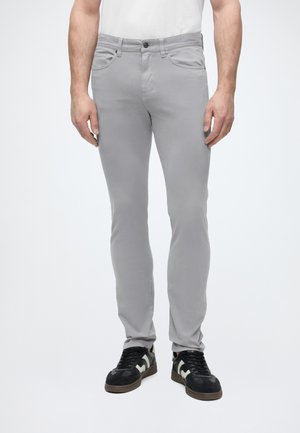 Férfi világosszürke slim fit nadrágban és fekete, fehér részletekkel díszített tornacipőben, egy egyszerű fehér háttér előtt áll.