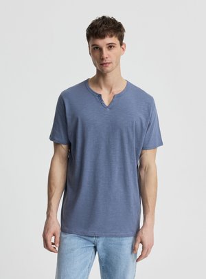 Giovane uomo che indossa una maglietta Henley blu a maniche corte e jeans azzurri, in piedi davanti a uno sfondo bianco semplice.