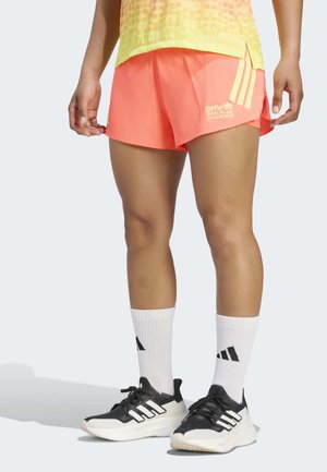 Piernas con pantalones cortos de running coral brillantes con el logo del Maratón de Berlín BMW, calcetines blancos de Adidas y zapatillas negras de running Adidas sobre un fondo claro.