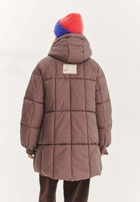 Brązowa kurtka puchowa z kapturem, pikowanym wzorem i widoczną metką na plecach. Do tego czerwona i niebieska dziana czapka.