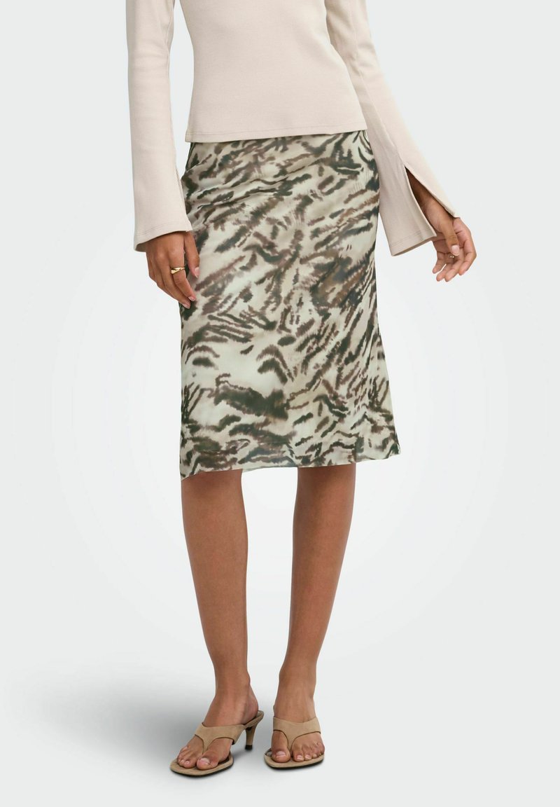 Femme portant un haut beige à manches longues fendues, une jupe mi-longue à motif animalier et des sandales beiges à talons, debout sur un fond uni.
