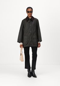 Dark green waterproof jacket with a brown collar, side pockets, and snap buttons, paired with black pants and a light handbag.