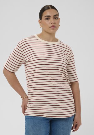 Młoda kobieta z ciemnymi włosami spiętymi do tyłu, nosząca złote kolczyki, kremowo-czerwoną pasiastą koszulkę, niebieskie dżinsy i złotą bransoletkę, pozuje neutralnie.