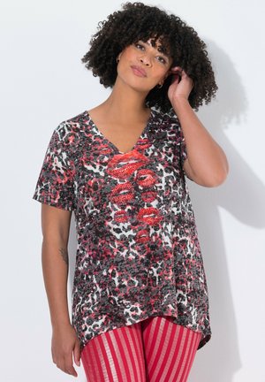 Femme aux cheveux bouclés portant un t-shirt à motifs rouge, noir et blanc avec un décolleté en V représentant des lèvres rouges, vêtu de pantalons rayés rouges et roses, touchant ses cheveux.