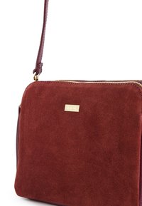Red suede crossbody bag with a textured surface, gold zipper detail, and a small logo plate. Adjustable strap for versatility.