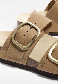 Sandales en daim marron avec deux larges brides, dotées d'une boucle en laiton et d'une semelle texturée marron. Semelle intérieure douce avec une finition naturelle.